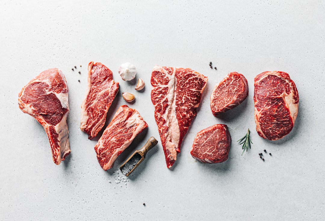 Verschiedene rohe Rindersteakstücke werden auf einer hellen Fläche mit einem Fleischbeil, Knoblauchzehen, Rosmarin, schwarzen Pfefferkörnern und Meersalz angerichtet. Das Fleisch ist marmoriert und hat eine leuchtend rote Farbe.